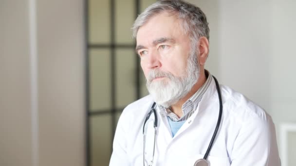 Bedachtzame zielige senior arts die naar binnen kijkt. Portret van een grijsharige blanke man die naar camera kijkt en handen kruist. Wanhoop en frustratie van medisch personeel bij een coronaviruspandemie. — Stockvideo