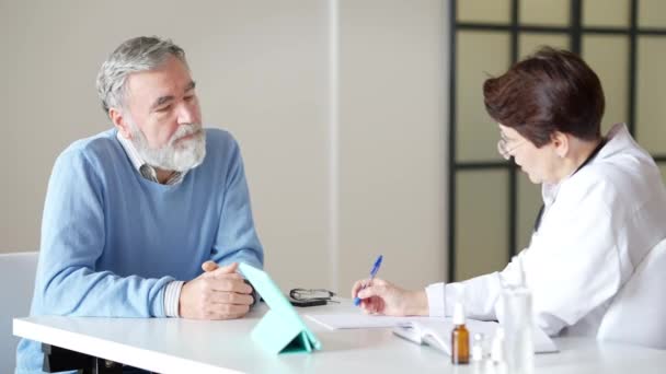 Paciente masculino mayor sentado en el consultorio médico hablando con una terapeuta. Retrato de un hombre caucásico barbudo de pelo gris que consulta a un médico en el hospital interior. Consulta de asistencia sanitaria y — Vídeos de Stock