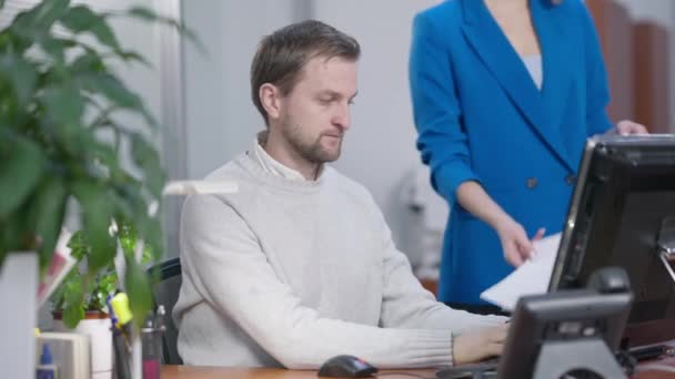 Ritratto di uomo caucasico impegnato che guarda attraverso documenti con donna irriconoscibile in piedi con le mani incrociate sullo sfondo. Maschio dipendente progetto di analisi per il capo femminile in ufficio. — Video Stock