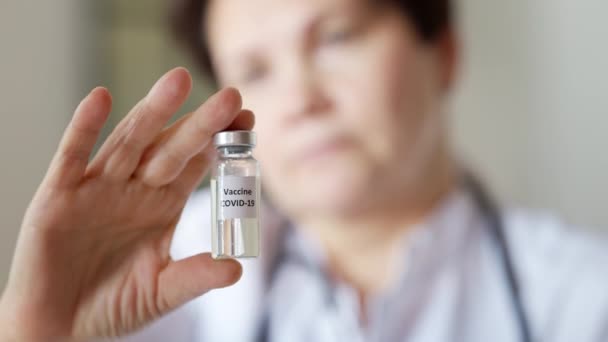 Gros plan du pot de vaccin contre le coronavirus dans la main blanche féminine avec le visage flou d'une femme grave d'âge moyen à l'arrière-plan. Médecin scientifique professionnel montrant le traitement Covid-19 à l'intérieur. — Video