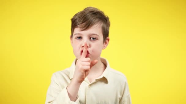 Portrait of serious boy putting finger at mouth showing hush gesture looking at camera. Brunette Caucasian child posing at yellow background asking for silence. — Stock Video