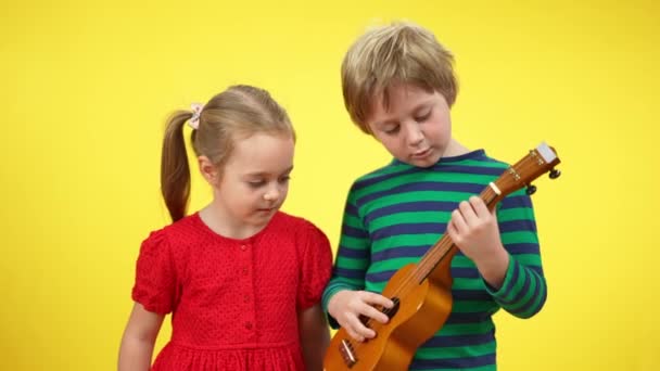 Bájos fehér testvér és húga játszik ukulele sárga háttér. Portré pozitív játékos gyerekek hangszer beszél és mosolyog. Hobbi és életmód. — Stock videók