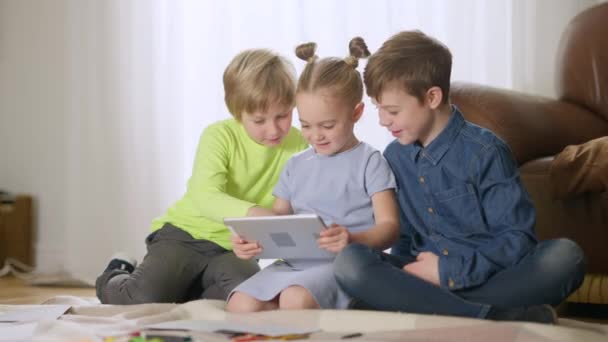 Трое очаровательных счастливых кавказских детей с помощью планшета разговаривают и улыбаются сидя в гостиной. Мальчики и девочки наслаждаются отдыхом вместе дома. Братья и сёстры смотрят мультфильмы онлайн в помещении. — стоковое видео