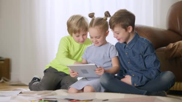 Tres encantadores niños caucásicos felices usando tabletas hablando y sonriendo sentados en la sala de estar. Positivos chicos y chicas disfrutando del ocio juntos en casa. Hermanos viendo dibujos animados online en interiores. — Vídeos de Stock