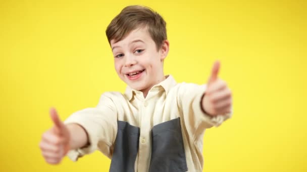 Opgewonden brunette jongen met duimen omhoog kijkend naar de camera en glimlachend naar gele achtergrond. Portret van vrolijk kind poseren op gekleurde achtergrond gebaren. — Stockvideo