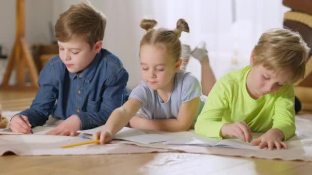 Koncentrált testvérek rajz festmény feküdt puha szőnyegen a nappaliban. Három pozitív kaukázusi gyerek pihen otthon a hétvégén. Egység és gyermekkori koncepció. — Stock videók