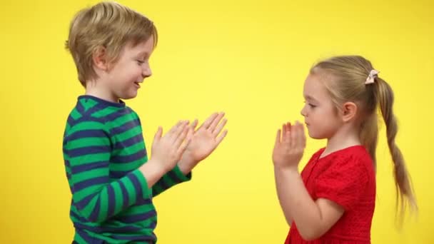 Vista lateral de niño y niña sin preocupaciones emocionados jugando juego de palmadas de mano en el fondo amarillo. Feliz hermano caucásico y hermana o amigos disfrutando de jugar y sonreír. Ocio infantil. — Vídeos de Stock