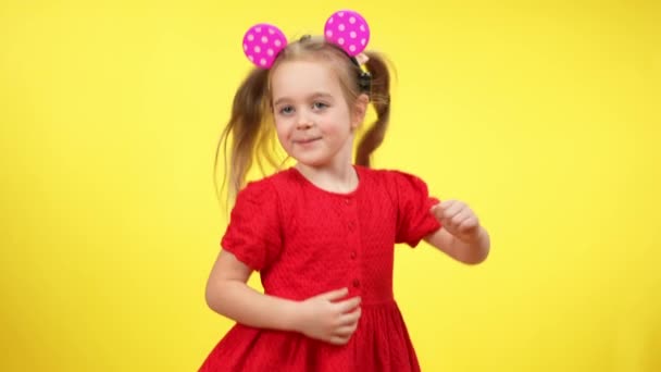 Linda chica alegre en vestido rojo y orejas de ratón de juguete bailando en cámara lenta en el fondo amarillo. Feliz niño caucásico alegre divirtiéndose. Felicidad y ocio infantil. — Vídeos de Stock