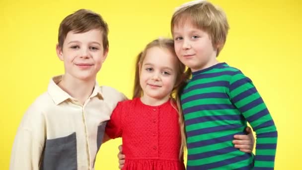 Three charming children hugging at yellow background and smiling looking at camera. Portrait of friends or siblings posing. Brothers and sister enjoying time together. Unity and family. — Stock Video