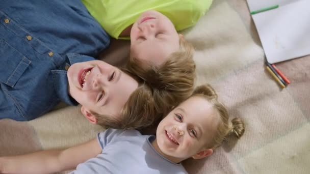 Зверху зображено трьох щасливих кавказьких дітей, які лежать на килимі, сміючись, дивлячись на камеру. Радісні брати і сестри або друзі, які добре відпочивають вдома. Дружба і дитяча концепція. — стокове відео