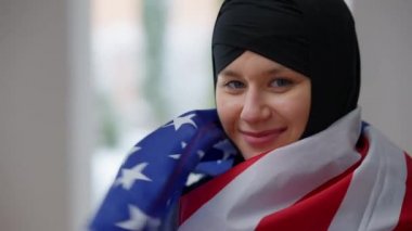 Amerikan bayrağına sarılmış, kameraya bakıp gülümseyen mutlu Müslüman kadının yakın çekimi. ABD ulusal sembolüyle içeride poz veren tatmin olmuş Orta Doğulu göçmen portresi. Göçmenlik kavramı