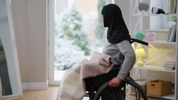 Zdravotně postižená mladá žena v hidžábu převaluje invalidní vozík ke skleněným dveřím. Paralyzoval Blízký východ krásné invalidní tráví osamělý den doma. Koncept zdravotního postižení a zvláštních potřeb. — Stock video