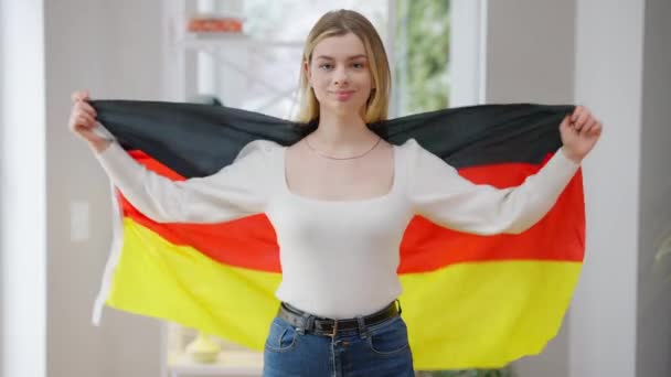 Retrato de jovem mulher confiante embrulhando em bandeira alemã olhando para a câmera sorrindo. Feliz senhora caucasiana relaxado posando com símbolo nacional em casa dentro de casa. Conceito de orgulho e patriotismo. — Vídeo de Stock