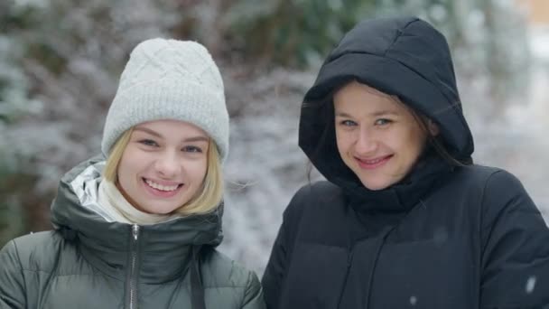 Close-up portret van twee vrolijke jonge vrouwen die poseren in de sneeuw in het dennenbos. Charmant mooi blank vrouwelijke vrienden genieten van ijzige winterdag buiten kijken naar camera glimlachen. — Stockvideo