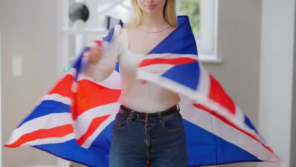 Joven esbelta irreconocible mujer británica envuelta en bandera nacional y sonriente de pie en casa. Alegre orgullosa dama caucásica posando en interiores. Inmigración y estilo de vida patriótico. — Vídeo de stock