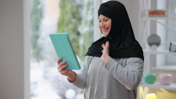 Joven mujer musulmana alegre en hijab ondeando en video chat sosteniendo la tableta, haciendo expresión facial sorprendida y riendo. tiro medio de mensajería de dama moderna segura positiva en línea. — Vídeos de Stock