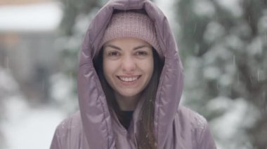 Kahverengi gözlü, dişlek gülüşlü, kar yağarken kameraya bakan neşeli esmer kadın portresi. Güzel, ince, mutlu, beyaz bir bayanın kışın açık havada dinlenen yüzü..