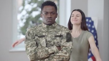 Elini çapraz yapıp kameraya bakan kendine güvenen bir askerin portresi gururlu bir kadının partnerine sarılması ve gülümsemesi. Afro-Amerikalı koca ve Kafkasyalı eş evde poz veriyor..
