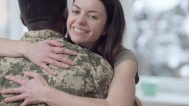 Afro-Amerikan askerini kucaklayan mutlu, gülümseyen beyaz kadın portresi kameraya bakıyor. Neşeli güzel eş eve dönen kocayı kucaklıyor. Aile ve mutluluk.