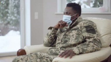 Covid-19 yüz maskeli ve askeri kamuflajlı genç Afro-Amerikan bir adamın yan görüntüsü. Koronavirüs salgınında evde dinlenen düşünceli aceminin portresi..