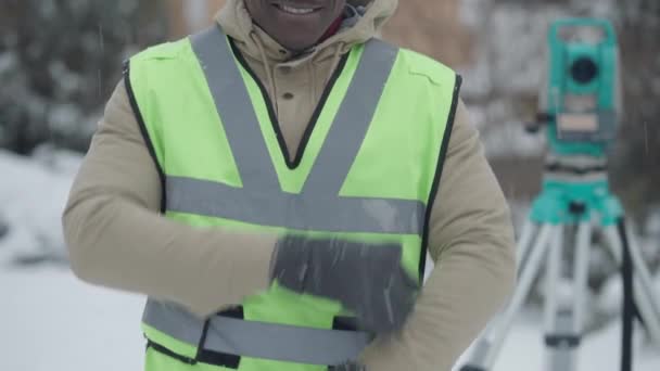 Felismerhetetlen mosolygós afro-amerikai férfi keresztezi egymás kezét elmosódott ipari felszereléssel teodolit a szabadban. Magabiztos fiatal geodézista pózol a téli napon. — Stock videók