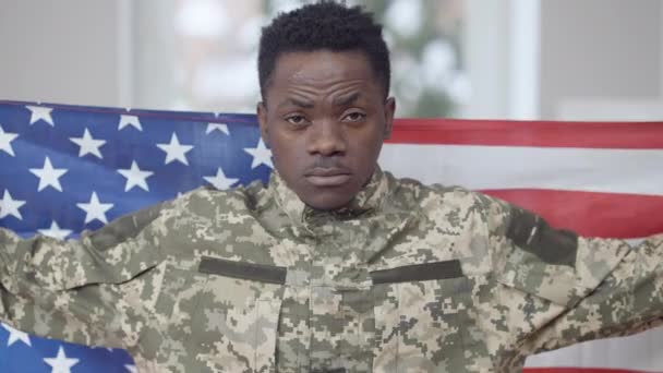 Retrato de jovem orgulhoso e confiante militar envolto em bandeira americana olhando para a câmera e sorrindo. Soldado afro-americano feliz posando com símbolo nacional dentro de casa. Conceito de patriotismo. — Vídeo de Stock