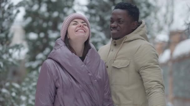 Fröhliches, fröhliches Paar, das im Schneefall steht, sich umarmt und lächelnd in die Kamera blickt. Porträt eines positiven afroamerikanischen hübschen Freundes und einer schönen kaukasischen Freundin, die draußen posiert. — Stockvideo