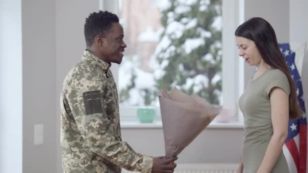 Vista laterale di amare l'uomo militare afroamericano dando mazzo di fiori per dimagrire bella donna caucasica. Felice eccitato giovane moglie incontro marito a casa al chiuso. Concetto di riunione. — Video Stock