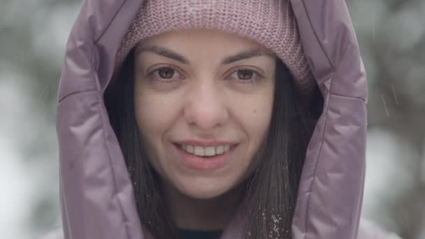Närbild ansikte säker vacker brunett kvinna med bruna ögon och toothy leende tittar på kameran som snö faller utomhus. Huvudskott av lycklig bekymmerslös kaukasisk turist på vintern resort. — Stockvideo