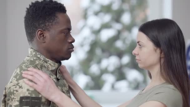 Visão lateral da triste jovem mulher caucasiana abraçando o marido afro-americano em uniforme militar e pensamento. Retrato de mulher bonita perturbada abraçando o homem que parte. Indústria da defesa e família. — Vídeo de Stock