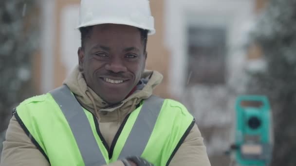 Close-up de confiante bonito homem afro-americano positivo em chapéu duro sorrindo olhando para a câmera ao ar livre. Retrato de geodésia agrimensor profissional posando no dia de inverno. Confiança engenharia. — Vídeo de Stock