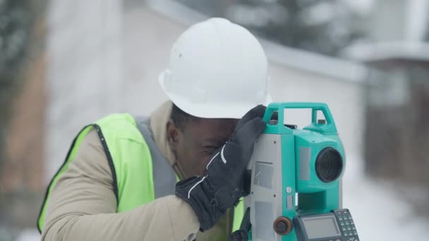 단단 한 모자를 쓴 자신감 있는 아프리카 계 미국인 측량사의 근접 사진. 눈내리는 겨울날에 일하는 젊고 잘생긴 남자. 직업 과 직업 개념. — 비디오