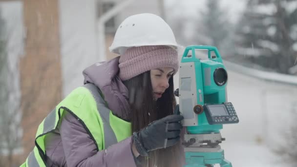 Soustředěná žena v klobouku s průmyslovým vybavením v zimě venku. Portrét profesionálního bělocha se zaměřením na geodézie pracujícího venku pomocí teodolitu. Koncept topografie. — Stock video