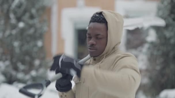 Porträt eines schwitzenden Afroamerikaners, der sich die Stirn wischt, nachdem er im Hinterhof Schnee gereinigt hat und in die Kamera blickt. Müder schöner junger Kerl posiert zu Hause im Garten mit Schaufel auf der Schulter. — Stockvideo