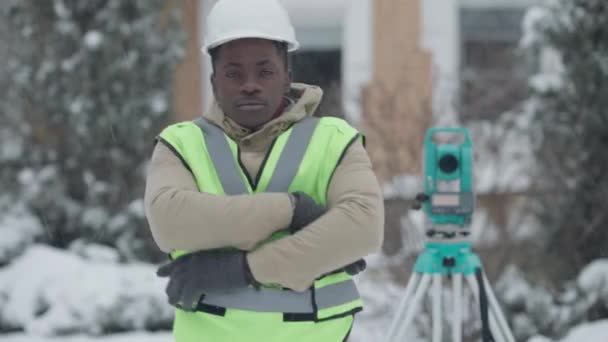 Középső lövés magabiztos pozitív profi földmérő kemény kalapban pózol szabadban Theodolite a háttérben. Egy afro-amerikai férfi kézen fogva nézi a mosolygó kamerát. Topográfia — Stock videók