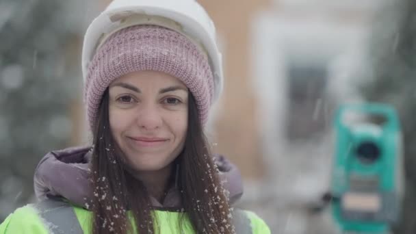 Close-up face of charming positive female topographer posing outdoors in snowfall. Portrait of beautiful young Caucasian woman in hard hat looking at camera smiling. Construction industry concept. — Stock Video