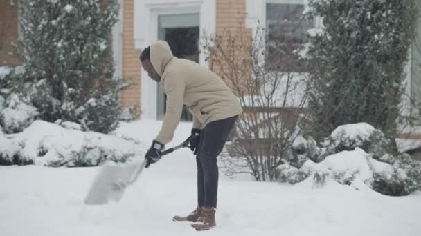 Amplo tiro de jovem afro-americano limpando neve sob queda de neve ao ar livre. Confiante concentrou cara bonito trabalhando no dia nevado com pá. Estilo de vida e conceito frio. — Vídeo de Stock