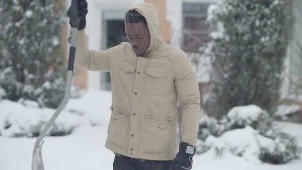 Colpo medio di uomo afroamericano sicuro in piedi con pala in nevicata. Ritratto di giovane ragazzo in posa all'aperto nella giornata invernale innevata. Pulizia neve di stagione fredda. — Video Stock