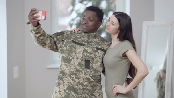 Gelukkig vrolijke interraciale paar nemen selfie op smartphone knuffelen. Vrolijke Afro-Amerikaanse man en blanke vrouw fotograferen thuis. Militaire man en vrouw die herinneringen vastleggen. — Stockvideo