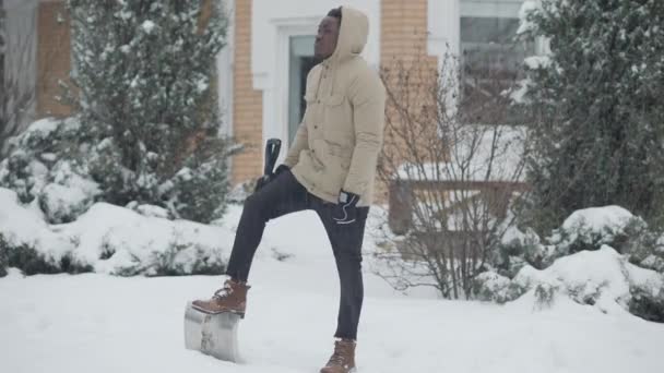 雪の冬の日に屋外でシャベルで立って思慮深いアフリカ系アメリカ人の男の広いショットの肖像画を見て回る。ハンサムな自信のある男クリーニング雪で家で庭. — ストック動画