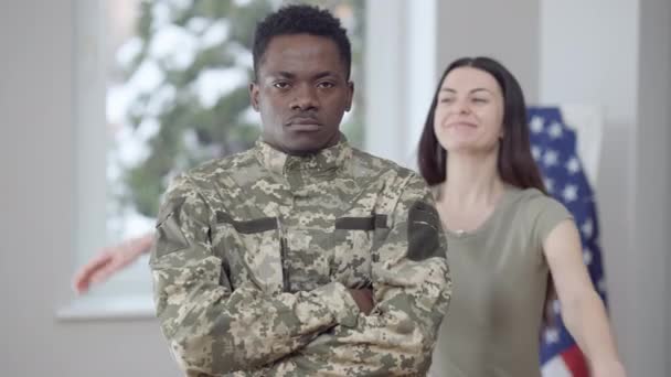 Portret van een zelfverzekerde militaire man met gekruiste handen kijkend naar de camera als trotse vrouw knuffelende partner en lachend. Afro-Amerikaanse man en blanke vrouw poseren binnen thuis. — Stockvideo
