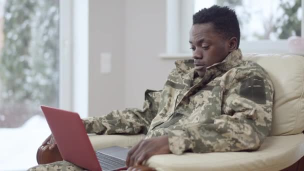 Självsäker ung militär man talar i videochatt sitter på fåtölj hemma. Porträtt av stilig afroamerikansk man i kamouflage uniform chatta online inomhus. Trådlös kommunikation. — Stockvideo