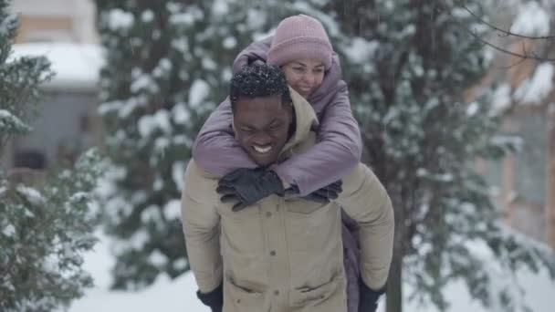 Positiv aussehender junger afroamerikanischer Mann, der sich dreht und eine charmante Kaukasierin auf dem Rücken hält. Porträt eines verliebten gemischtrassigen Paares, das den Winter im Freien genießt. Freizeit und Lebensstil. — Stockvideo