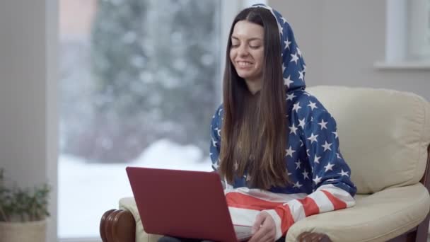 Positieve vrolijke jonge brunette vrouw in hoodie gekleurd aan Amerikaanse vlag praten in video chat en lachen. Portret van ontspannen zorgeloze blanke slanke dame genieten van draadloze communicatie binnen thuis — Stockvideo