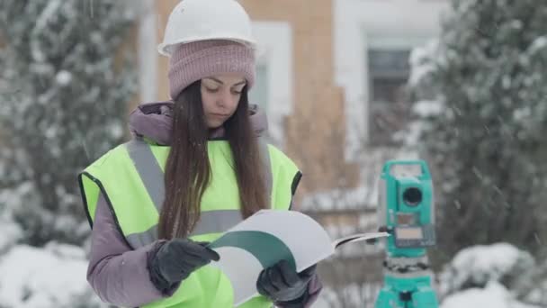 Koncentrerad kvinnlig arkitekt undersöker projektdokumentation på snöig vinterdag utomhus med teodolit i bakgrunden. Porträtt av professionell entreprenör kvinna som arbetar på byggarbetsplatsen. — Stockvideo