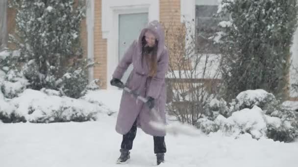 Vreugdevolle slanke charmante vrouw die sneeuw schoonmaakt met schep en buiten lacht. Breed shot portret van vrolijke blanke dame genieten van sneeuw op de achtertuin. Vrije tijd en levensstijl. — Stockvideo