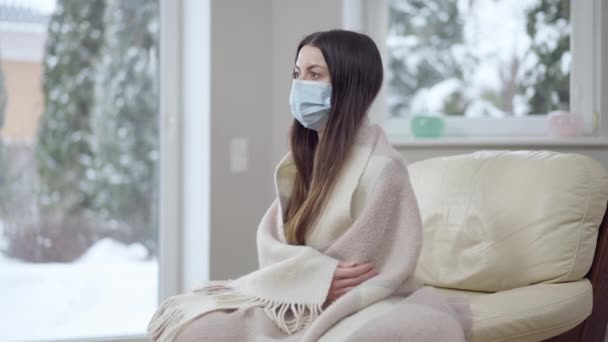 Verdrietige jonge zieke vrouw in coronavirus gezichtsmasker zittend op fauteuil verpakt in een deken die wegkijkt. Zijaanzicht portret van jonge blanke dame lijdt aan symptomen van Covid-19 pandemische ziekte. — Stockvideo