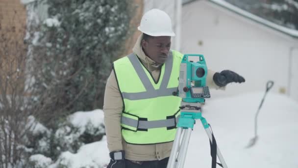 Profesionální sebevědomý geodézista používající teodolit venku za sněhového zimního dne. Střední snímek seriózního afroamerického pohledného muže v klobouku. Geodézie a inženýrství. — Stock video