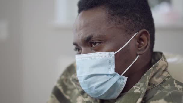 Närbild ansikte ledsen afroamerikansk man i coronavirus ansikte mask tittar bort tänkande inomhus. Huvudskott porträtt av stilig ung soldat inomhus på Covid-19 pandemi utbrott. — Stockvideo