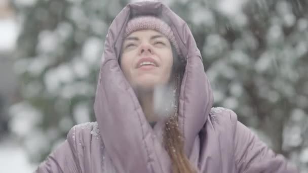 Zbliżenie portret szczęśliwej, szczupłej młodej białej kobiety rzucającej śniegiem i uśmiechającej się patrząc w kamerę. Radosny, relaksujący, beztroski tysiącletnie zabawy na świeżym powietrzu w mroźny zimowy dzień. Koncepcja czasu wolnego. — Wideo stockowe